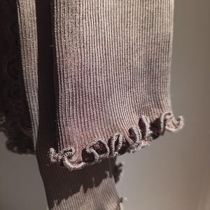 K&ouml;nnte beinhalten: Nahaufnahme eines braunen Rippstrickstoffs mit R&uuml;schenkante. Der Stoff hat eine vertikale Rippstruktur und ist einfarbig braun. Der R&uuml;schenrand verleiht dem Kleidungsst&uuml;ck ein dekoratives Detail.