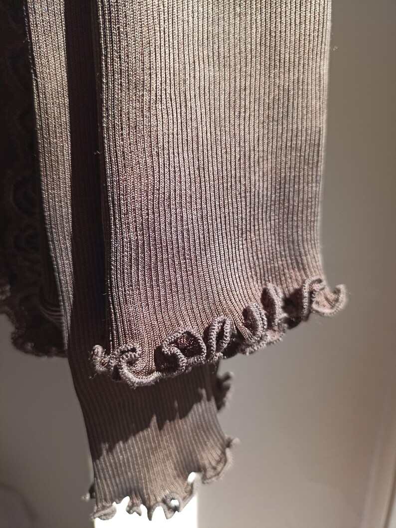 K&ouml;nnte beinhalten: Nahaufnahme eines braunen Rippstrickstoffs mit R&uuml;schenkante. Der Stoff hat eine vertikale Rippstruktur und ist einfarbig braun. Der R&uuml;schenrand verleiht dem Kleidungsst&uuml;ck ein dekoratives Detail.