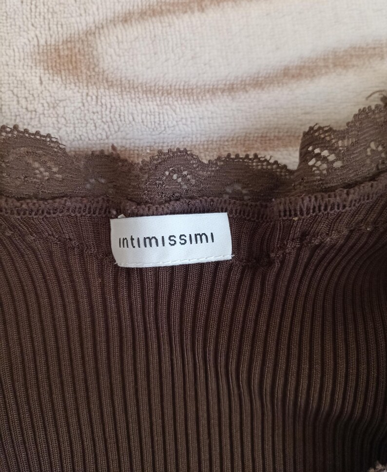 K&ouml;nnte beinhalten: Nahaufnahme eines braunen Ripp-Tanktops mit Spitzenbesatz. Auf dem Etikett steht "intimissimi" in einem wei&szlig;en Rechteck. Das Oberteil besteht aus einem weichen, gerippten Material.