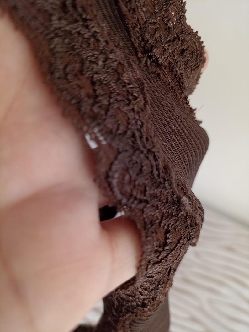 K&ouml;nnte beinhalten: Nahaufnahme eines braunen Kleidungsst&uuml;cks mit einem strukturierten, spitzenartigen Besatz. Der Stoff scheint ein dunkler Braunton zu sein, mit einem gerippten Abschnitt. Das Bild hebt die komplizierten Details des Textils hervor.