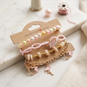 Può includere: Un set di quattro braccialetti fatti a mano su una scheda marrone con il testo "Handmade with love". I braccialetti presentano perle, perline rosa, catena dorata e ciondoli, tra cui cuori e un fenicottero. Un ditale e una bobina di filo sono sullo sfondo.