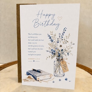 May include: A white birthday greeting card featuring blue text that reads Happy Birthday, a Bible verse from Numbers 6:24-26, and an illustration of a glass jar filled with blue and white wildflowers next to a closed book with a pen on top.