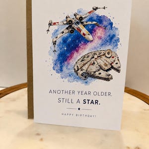 May include: A white greeting card featuring a watercolor illustration of an X-wing starfighter and the Millennium Falcon against a blue and purple nebula. The text reads: ANOTHER YEAR OLDER. STILL A STAR. HAPPY BIRTHDAY! with a small star icon.