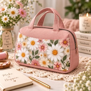 May include: A dusty pink fabric handbag featuring a front panel with a white and pink daisy floral pattern. The bag has two top handles, a zippered closure, and a structured rectangular shape with rounded corners and gold-toned hardware.