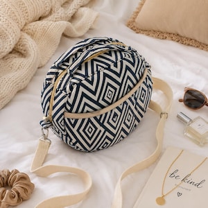 May include: A round crossbody bag featuring a navy blue and white geometric diamond pattern. The bag has a gold-tone zipper closure, a top handle, and a long, adjustable cream-colored canvas strap. The front includes a curved slip pocket.