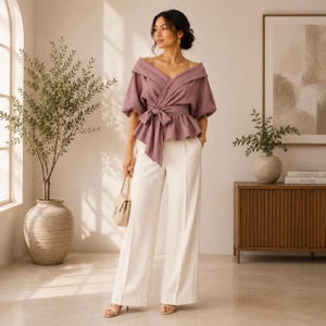 Può includere: Una camicetta a portafoglio color malva con ampio colletto ripiegato, maniche a sbuffo fino al gomito e cintura in vita, abbinata a pantaloni color crema a vita alta e gamba larga con cucitura centrale e una piccola borsa beige con catena dorata.