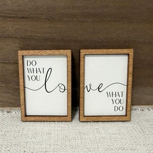 May include: Two rectangular wooden signs with white backgrounds and black text. The left sign reads DO WHAT YOU, and the right sign reads WHAT YOU DO, with the word LOVE written in a cursive script across both signs connecting the two phrases.