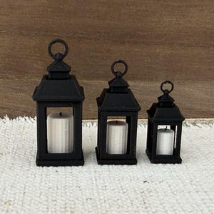 May include: Three black metal lanterns of varying sizes arranged in a row. Each lantern features a square frame, a top hanging loop, and a single white cylindrical pillar candle placed inside the central glass-paneled compartment.
