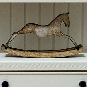 May include: A miniature wooden rocking horse ornament featuring a distressed white and natural wood finish. The horse is depicted in a galloping pose, mounted on a curved base that allows for a rocking motion, showcasing visible wood grain textures.