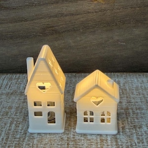 May include: Two white ceramic house-shaped tea light holders with illuminated interiors. The taller house features a chimney and heart-shaped cutout, while the shorter house has a heart-shaped cutout and multiple rectangular window openings.