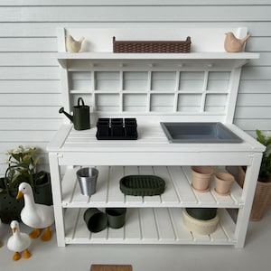 May include: A white wooden potting bench with a slatted work surface, a gray sink, and two lower shelves. Accessories include a dark green watering can, black seedling trays, small pots, a brown woven basket, and three white goose figurines.