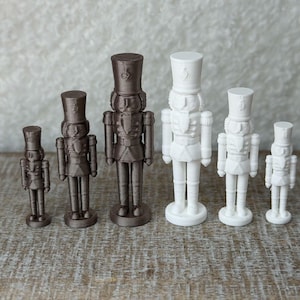 May include: A collection of six standing nutcracker figurines arranged in a row. Three are dark brown and three are white. Each nutcracker features a tall hat, a uniform with detailed buttons, and a circular base. The figures vary in height.