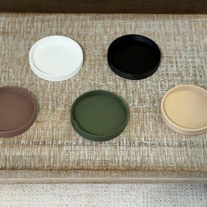 May include: Five circular, shallow trays arranged in a group. The set includes one white, one black, one brown, one olive green, and one beige tray. Each tray features a smooth, solid-colored surface with a raised rim, providing a simple, flat design.