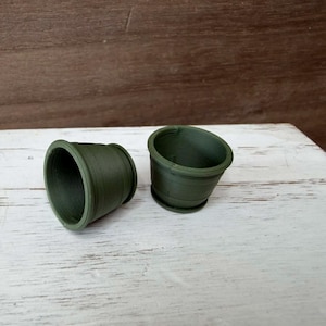 May include: Two small, olive green plant pots are displayed on a white wooden surface. One pot is upright, while the other is on its side. The pots have a simple, rounded design, suitable for small plants or succulents.