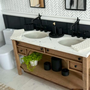 May include: Miniature bathroom vanity with two sinks, black faucets, and a white countertop. The wooden cabinet has drawers and a lower shelf with decorative items. A small potted plant adds a touch of greenery. The background features a patterned wall and a toilet.