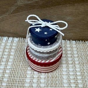 May include: A stack of four small, round boxes tied with white string. The top box is navy blue with white stars. The next box is white with a scalloped edge. The third box is gray, and the bottom box is red and white striped.