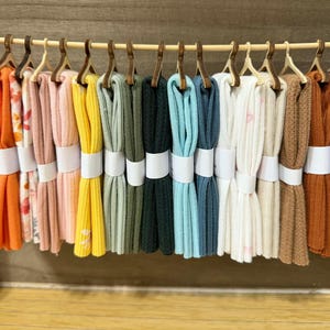 May include: A row of fifteen folded fabric swatches in various colors including red, orange, yellow, green, blue, white, and brown, each hanging from a small wooden hanger on a horizontal rod, secured with a plain white paper band around the center.