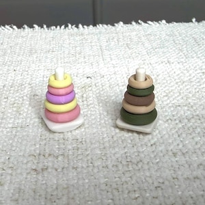 May include: Two miniature stacking ring toys on white square bases. The left toy features rings in pastel yellow, pink, and lavender. The right toy features rings in shades of olive green, brown, and tan. Both have a central white post.