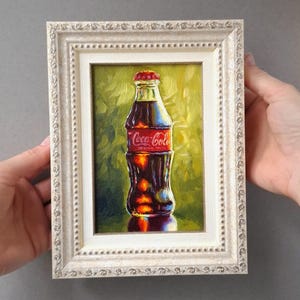 Peut inclure: Peinture à l'huile d'une bouteille de Coca-Cola dans un cadre blanc orné. La bouteille est brun foncé avec une étiquette rouge et un bouchon rouge. Le fond est un mélange de teintes vertes et jaunes.