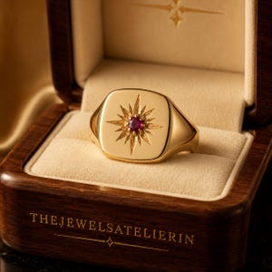 Peut inclure: Une bague chevalière en or ornée d'une pierre précieuse rouge centrale entourée d'un motif d'étoile gravé. La bague est présentée dans une boîte en bois avec le texte THEJEWELSATELIERIN imprimé en doré sur le bord avant du couvercle.