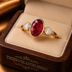 May include: A gold ring featuring a large oval-cut red gemstone in a bezel setting, flanked by two smaller pear-shaped white diamonds. The ring is presented in a wooden box with the text THEJEWELSATELIERIN engraved on the front edge.