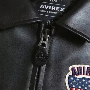 以下が含まれることがあります： 黒い革のジャケットのクローズアップ。ジッパー開閉式。赤、白、青の色を使ったパッチが付いています。「AVIREX」のブランド名がジャケットのラベルに表示されています。