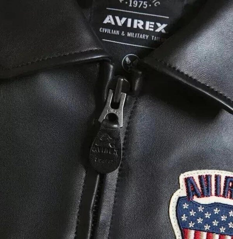 以下が含まれることがあります： 黒い革のジャケットのクローズアップ。ジッパー開閉式。赤、白、青の色を使ったパッチが付いています。「AVIREX」のブランド名がジャケットのラベルに表示されています。