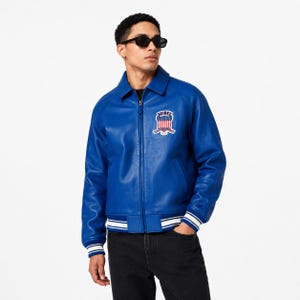 May include: A vibrant blue leather jacket with a zip-up front, a classic collar, and a patch on the chest. The jacket features white stripes on the cuffs and hem. The patch displays a patriotic emblem with the word "AVIREX".