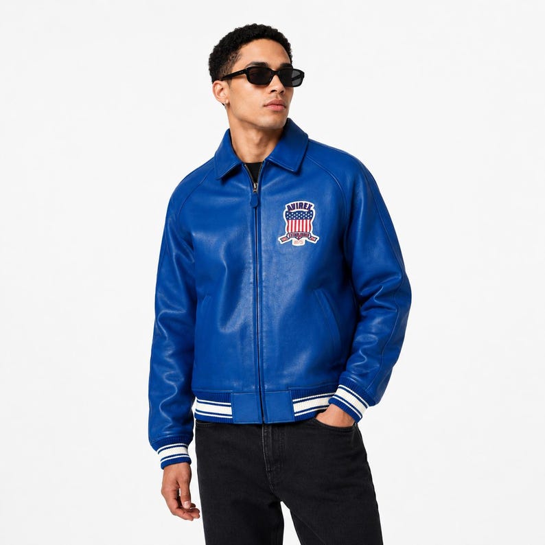 May include: A vibrant blue leather jacket with a zip-up front, a classic collar, and a patch on the chest. The jacket features white stripes on the cuffs and hem. The patch displays a patriotic emblem with the word "AVIREX".