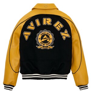 May include: A varsity-style jacket with a black body and mustard yellow leather sleeves. The back features the word "AVIREX" and a circular emblem with "SINCE 1975 NEW YORK CLASSIC" text. The jacket has a collar and striped cuffs.