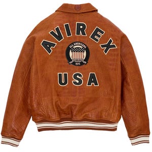May include: A brown leather bomber jacket with a crocodile-embossed texture. The back features the text AVIREX in black letters, a circular patch with an American flag and the text ALL AMERICANS ESTABLISHED 1975, and the text USA in black.