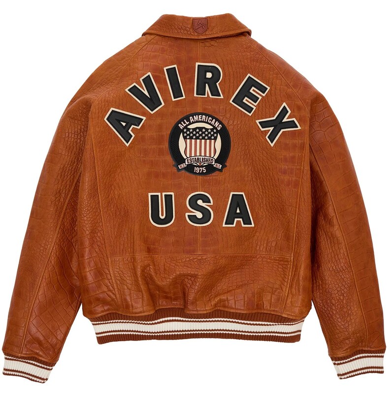 May include: A brown leather bomber jacket with a crocodile-embossed texture. The back features the text AVIREX in black letters, a circular patch with an American flag and the text ALL AMERICANS ESTABLISHED 1975, and the text USA in black.