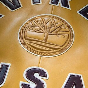 K&ouml;nnte beinhalten: Eine hellbraune Lederjacke mit einem kreisf&ouml;rmigen, gestickten Baumlogo in der Mitte. Das Design umfasst dunkelbraune und wei&szlig;e Blockbuchstaben, die USA um das Emblem bilden, mit sichtbaren N&auml;hten auf der glatten Lederoberfl&auml;che.