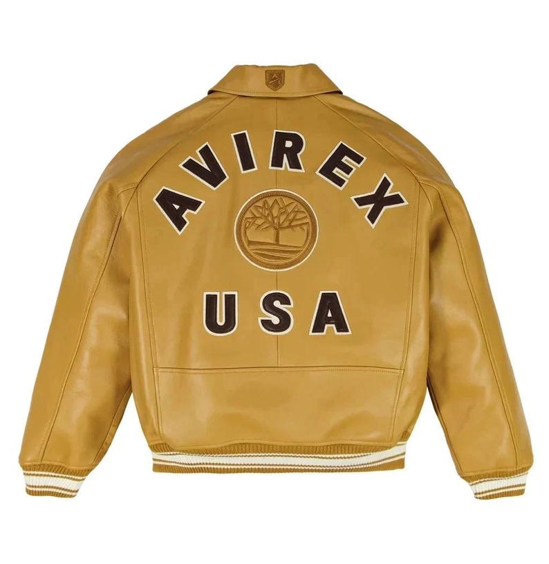 K&ouml;nnte beinhalten: Eine hellbraune Lederjacke mit Kragen und gerippten B&uuml;ndchen mit wei&szlig;en Streifen. Auf der R&uuml;ckseite befindet sich der Schriftzug AVIREX in einem dunkelbraunen Bogen, ein kreisf&ouml;rmiges Baum-Emblem in der Mitte und der Schriftzug USA darunter.