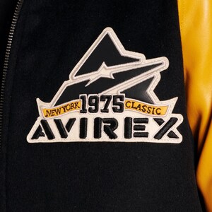 May include: Close-up of a black and yellow varsity jacket. The jacket features a patch with the text "NEW YORK 1975 CLASSIC AVIREX" in white, black, and yellow. The jacket has a black body and yellow sleeves.