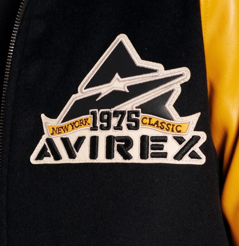 May include: Close-up of a black and yellow varsity jacket. The jacket features a patch with the text "NEW YORK 1975 CLASSIC AVIREX" in white, black, and yellow. The jacket has a black body and yellow sleeves.