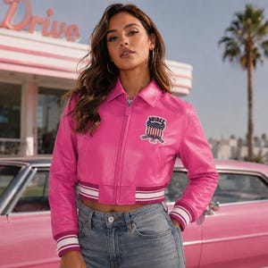 May include: A bright pink leather jacket with a zip-up front, a collar, and a patch on the chest. The jacket has white and burgundy striped cuffs and waistband. The model is wearing blue jeans. The background includes a pink car and a retro diner sign.