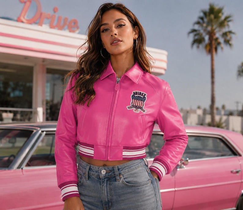 May include: A bright pink leather jacket with a zip-up front, a collar, and a patch on the chest. The jacket has white and burgundy striped cuffs and waistband. The model is wearing blue jeans. The background includes a pink car and a retro diner sign.