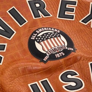 May include: A tan jacket with a crocodile-embossed texture featuring large black and white block letters and a circular patch. The patch displays an American flag shield, the text ALL AMERICANS ESTABLISHED 1975, and N.Y. in black and white.