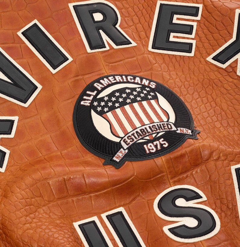 May include: A tan jacket with a crocodile-embossed texture featuring large black and white block letters and a circular patch. The patch displays an American flag shield, the text ALL AMERICANS ESTABLISHED 1975, and N.Y. in black and white.
