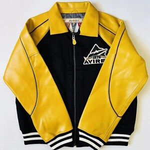 May include: A black and yellow jacket with a zip-up front. The jacket has yellow leather sleeves and collar, with black fabric on the body. The jacket has the text "AVIREX NEW YORK 1975 CLASSIC" on the chest and white stripes on the cuffs.