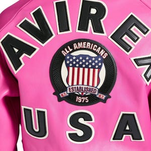 May include: Bright pink jacket with black and white lettering spelling out "AVIRE USA". A circular patch features an American flag design and the words "ALL AMERICANS ESTABLISHED 1975".