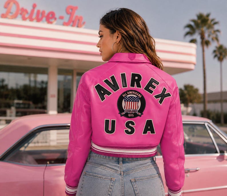 May include: A bright pink leather jacket with the word "AVIREX" in black lettering across the back, along with a circular patch featuring the American flag. The jacket has a ribbed hem and cuffs with white stripes. The background includes a pink car and a retro diner.