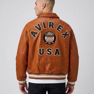 May include: A tan leather bomber jacket with a textured finish, featuring the text AVIREX in black letters across the upper back, a circular patch with a flag design, and the word USA in black letters below, with striped ribbed cuffs and hem.