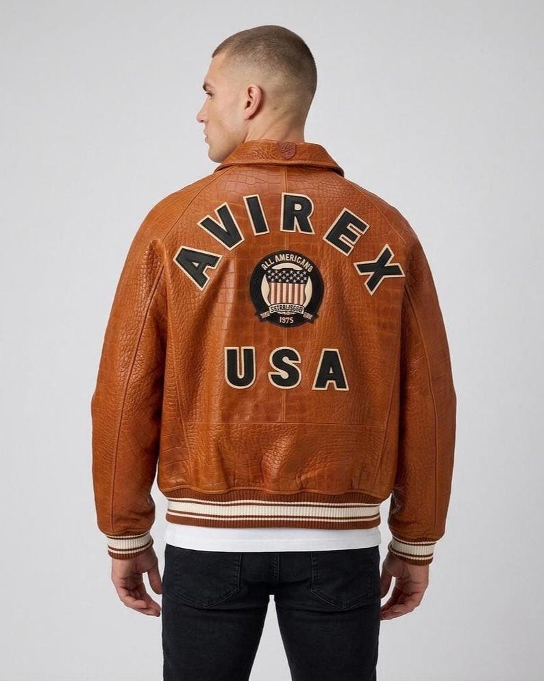 May include: A tan leather bomber jacket with a textured finish, featuring the text AVIREX in black letters across the upper back, a circular patch with a flag design, and the word USA in black letters below, with striped ribbed cuffs and hem.