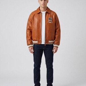 May include: A tan, crocodile-textured leather bomber jacket with a fold-down collar, ribbed cuffs and hem featuring white and brown stripes, and a circular patch on the left chest displaying the text "UNITED" above a shield graphic.
