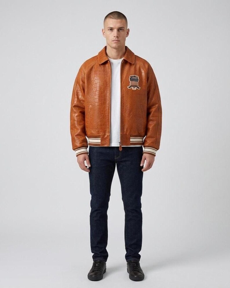 May include: A tan, crocodile-textured leather bomber jacket with a fold-down collar, ribbed cuffs and hem featuring white and brown stripes, and a circular patch on the left chest displaying the text "UNITED" above a shield graphic.