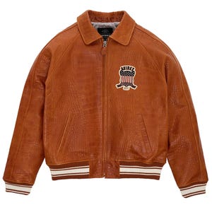 May include: A tan leather jacket with a crocodile-embossed texture, featuring a front zipper closure, a pointed collar, and ribbed cuffs and hem with white stripes. A patch on the chest displays the text AVIREX with a flag graphic.
