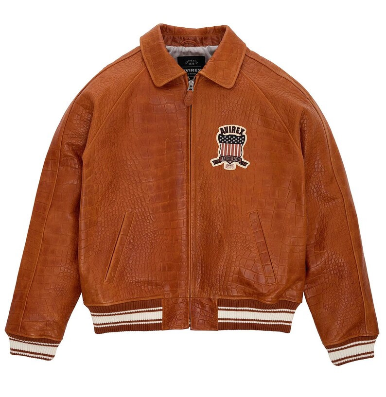May include: A tan leather jacket with a crocodile-embossed texture, featuring a front zipper closure, a pointed collar, and ribbed cuffs and hem with white stripes. A patch on the chest displays the text AVIREX with a flag graphic.
