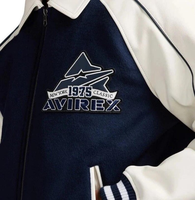 May include: A navy blue and white varsity jacket with a white collar and sleeves. The jacket features a patch with the text "NEW YORK 1975 CLASSIC AVIREX" in white and navy blue.